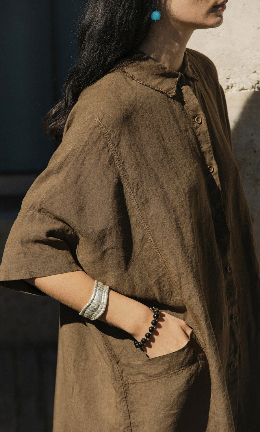 Plümo- Woman in the Hockley Linen Dress with a black bead bracelet, turquoise earring, hand in pocket, standing in sunlight.