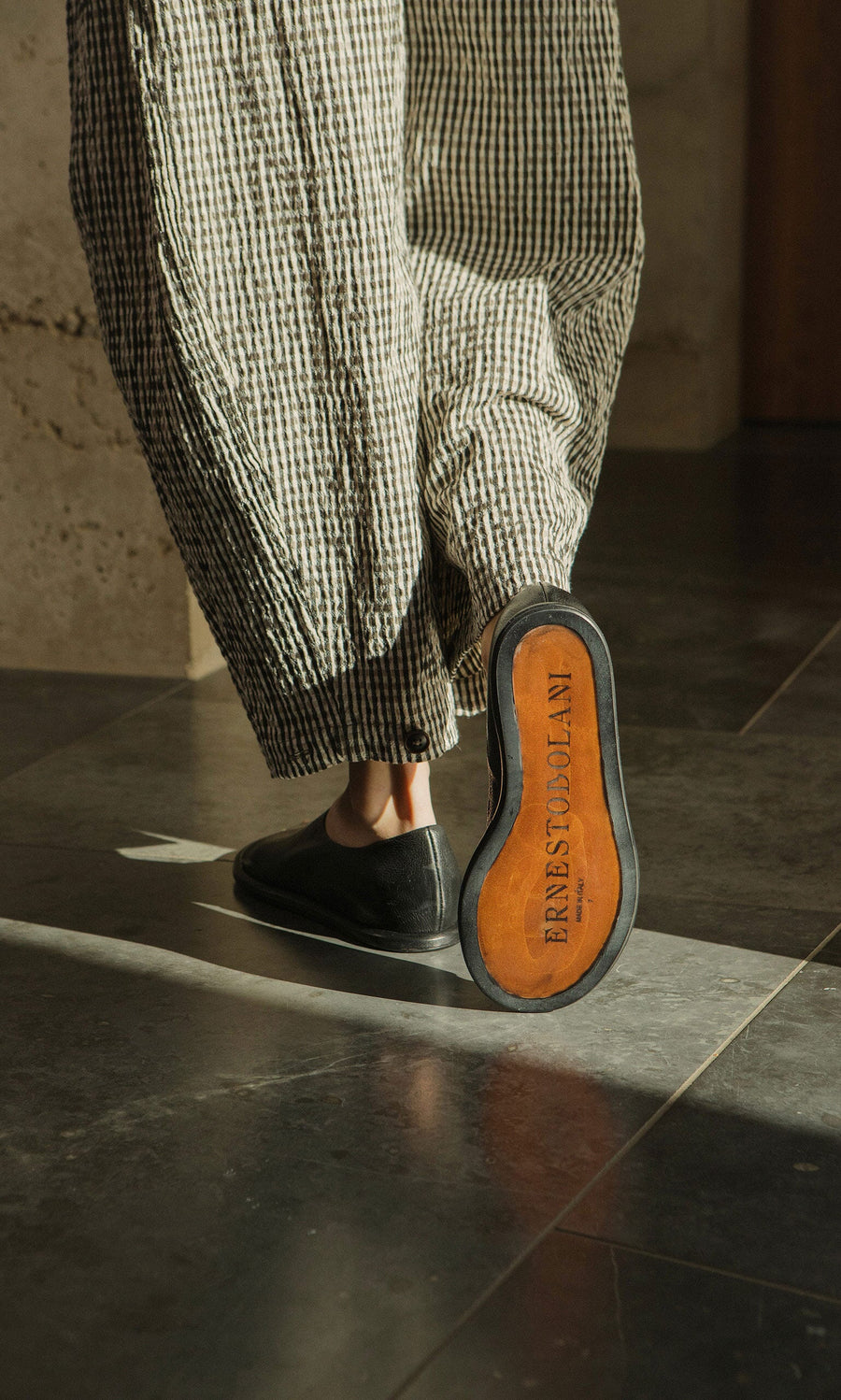 Plümo- Person in checkered pants wearing Hawke Sandals; sole reads FARFETCH X COLLAN as they walk on a tiled floor.