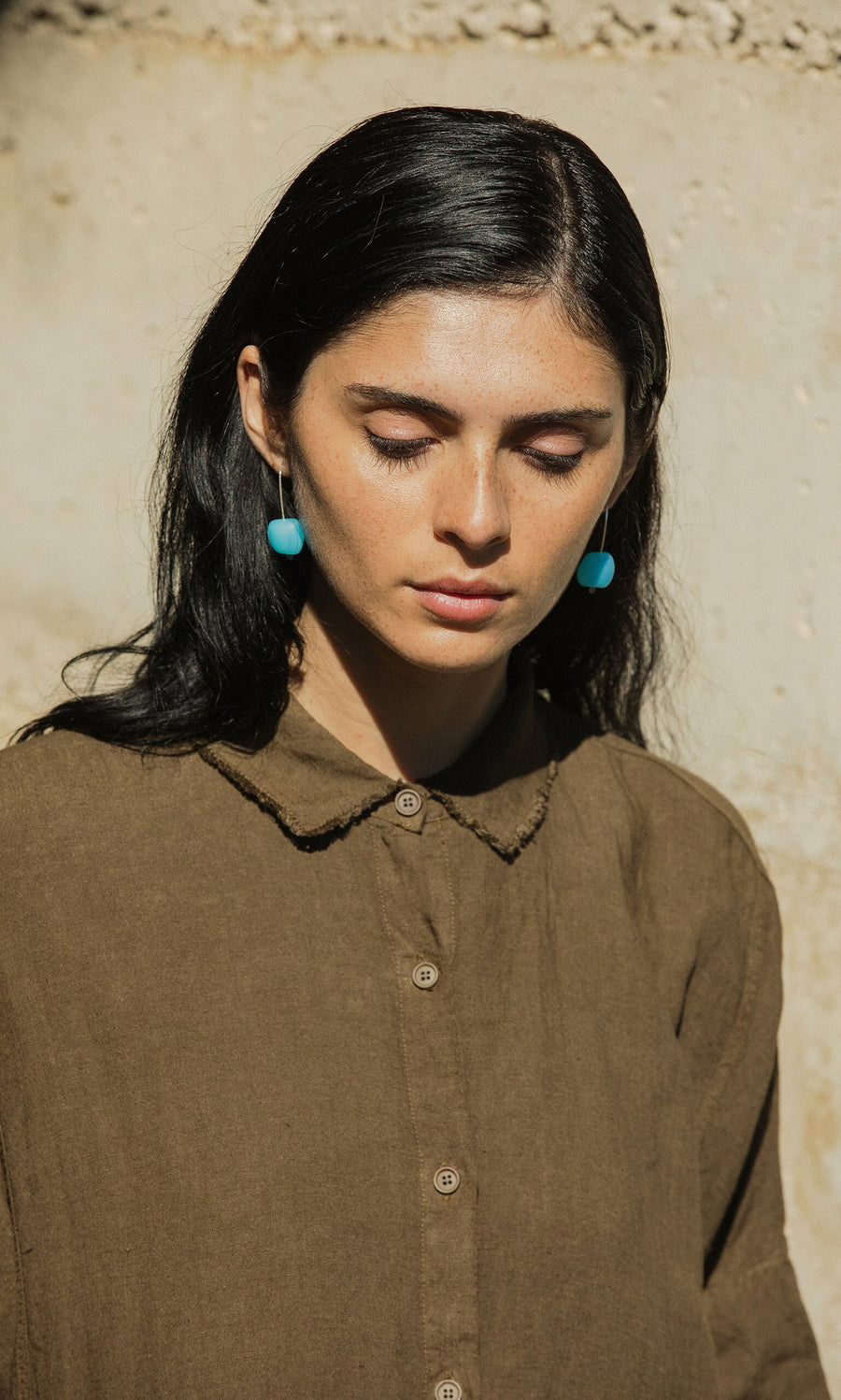 Plümo- Woman with long dark hair wears the Hockley Linen Dress and turquoise earrings, standing by a light-colored wall.