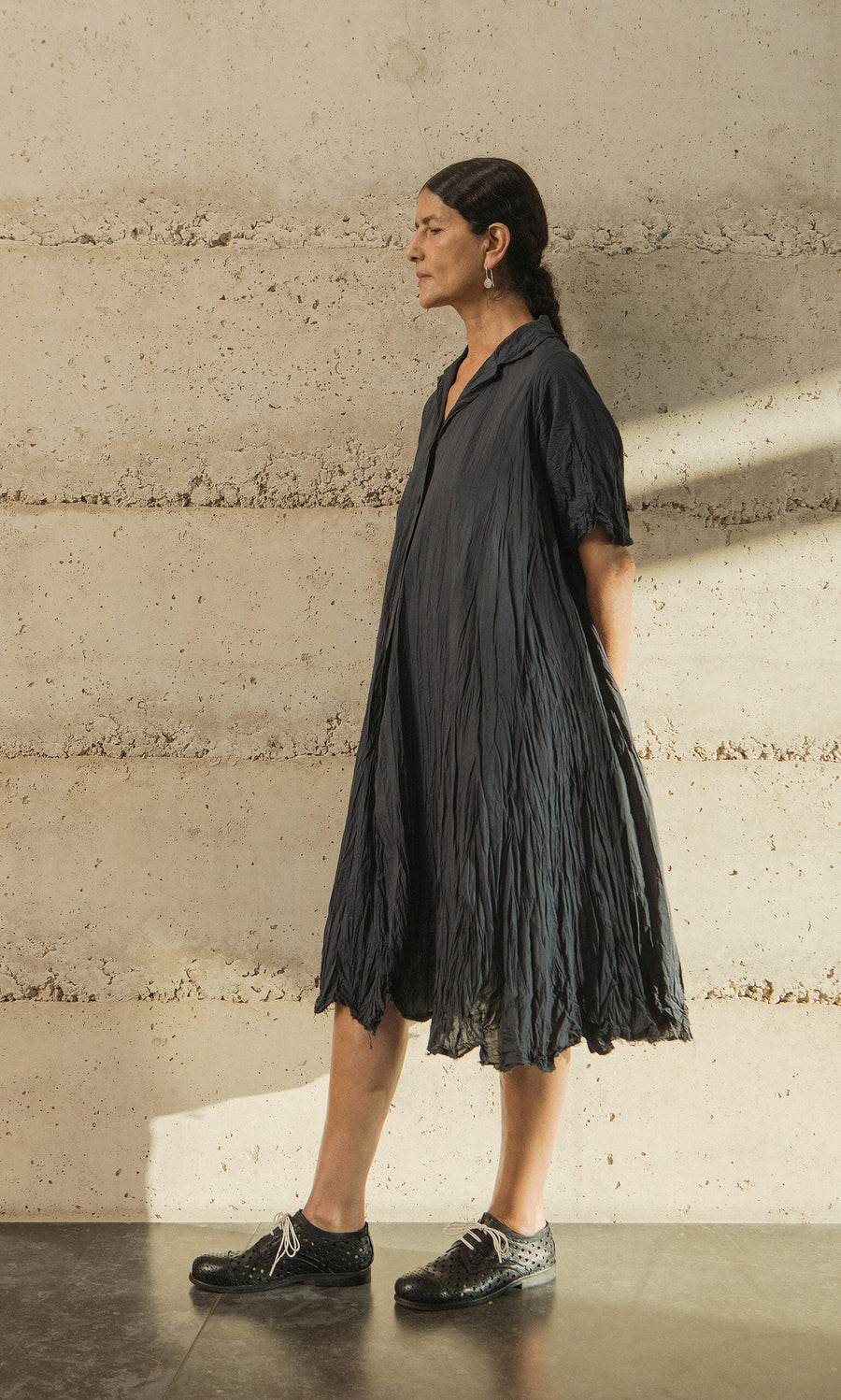 Plümo- Woman in the Brook Crinkle Dress and black shoes stands sideways against a textured beige wall.