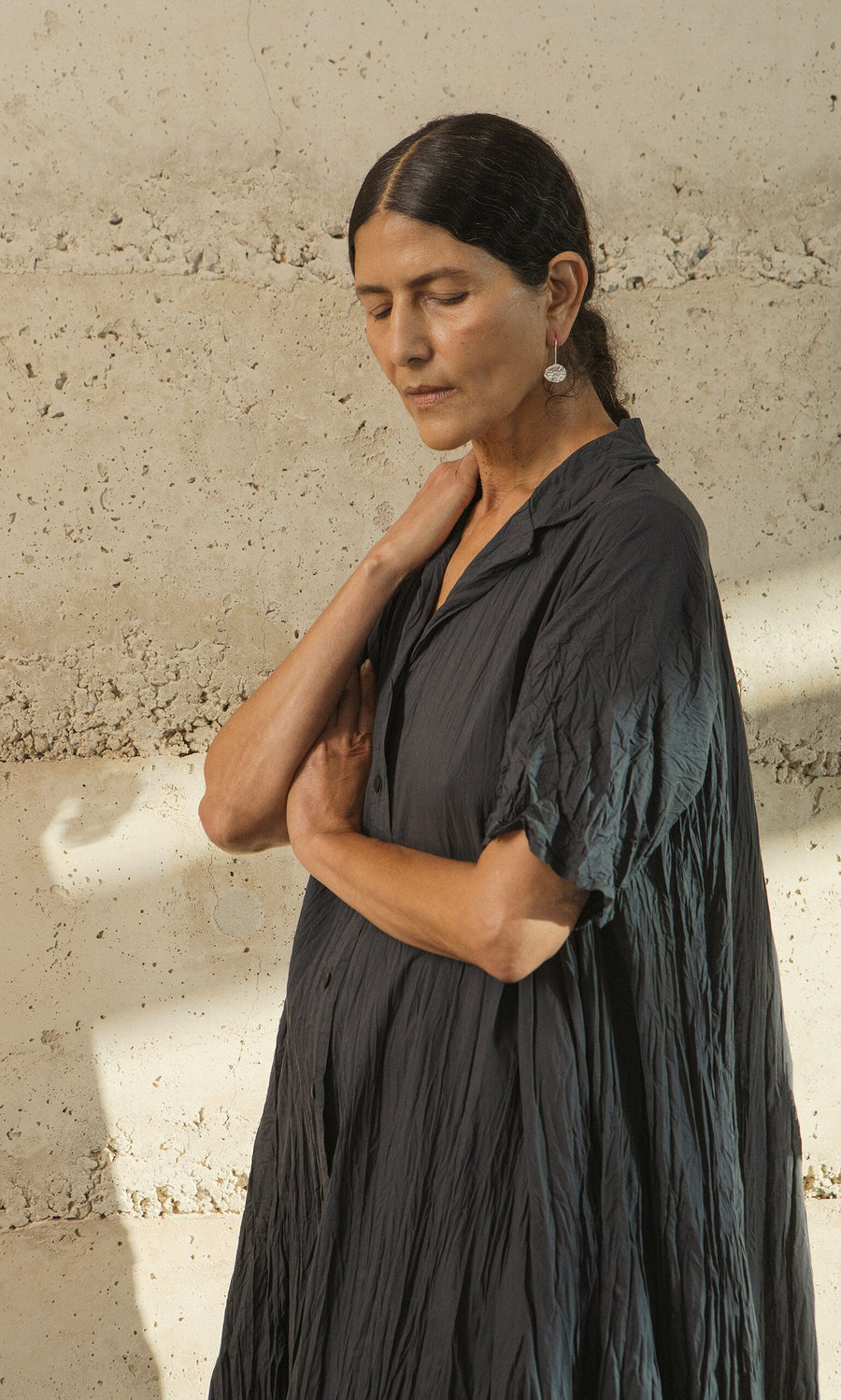 Plümo- A woman in the Brook Crinkle Dress stands against a textured wall, eyes closed, hand on her neck.