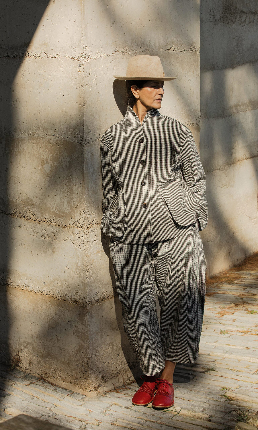 Plümo- Person in a striped suit, beige hat, and Magui Brogues leans against a concrete wall in dappled sunlight.