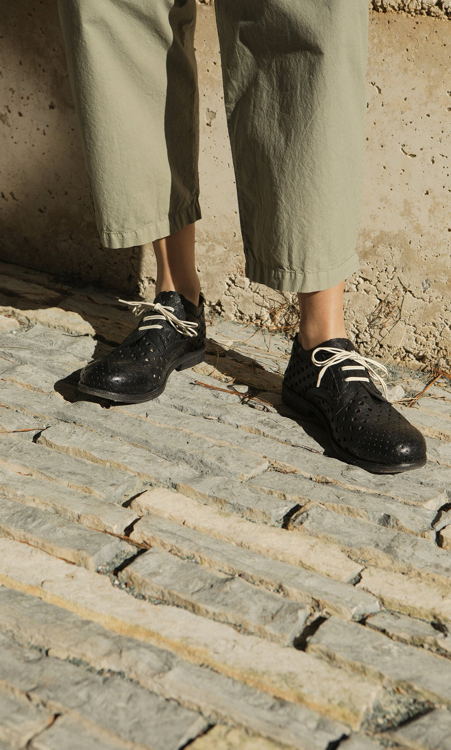 Plümo- Person wearing light green pants and Black Derby Brogues, standing on textured stone pavement in sunlight.