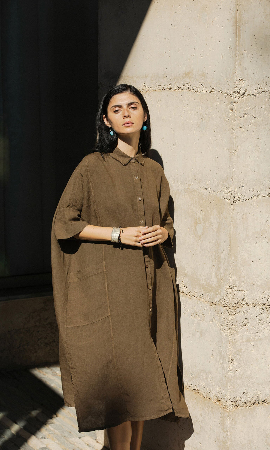Plümo- Woman in the Hockley Linen Dress with a raw edge collar stands calmly against a sunlit wall, looking forward.