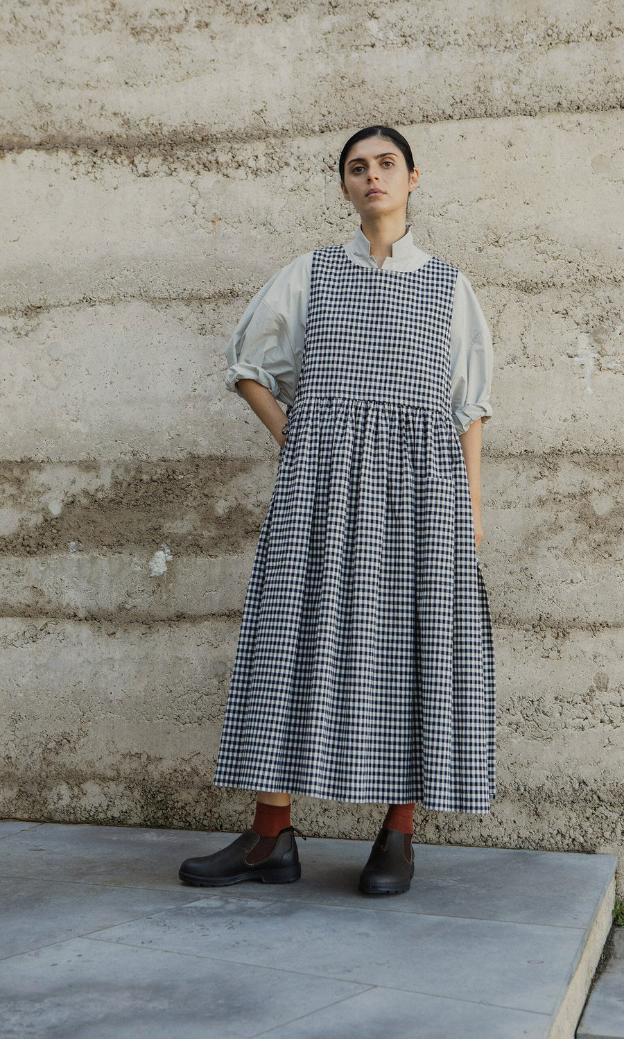 Plümo- A person in the Apron Dress - Gingham with spacious pockets stands against a textured wall, looking upward.