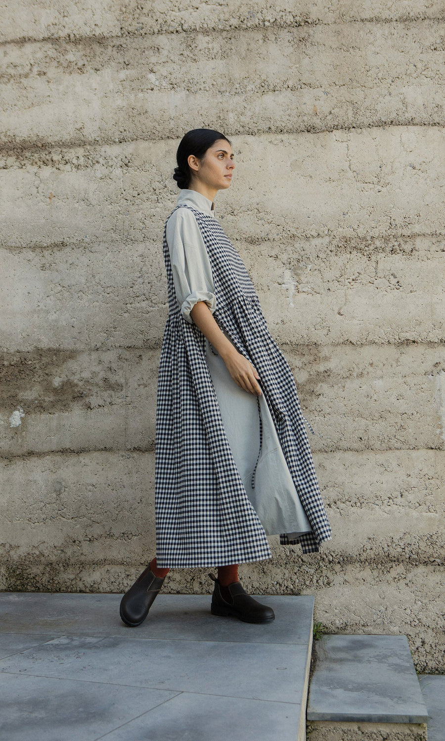 Plümo- A woman in the Apron Dress - Gingham and boots walks past a textured beige wall on a stone floor.