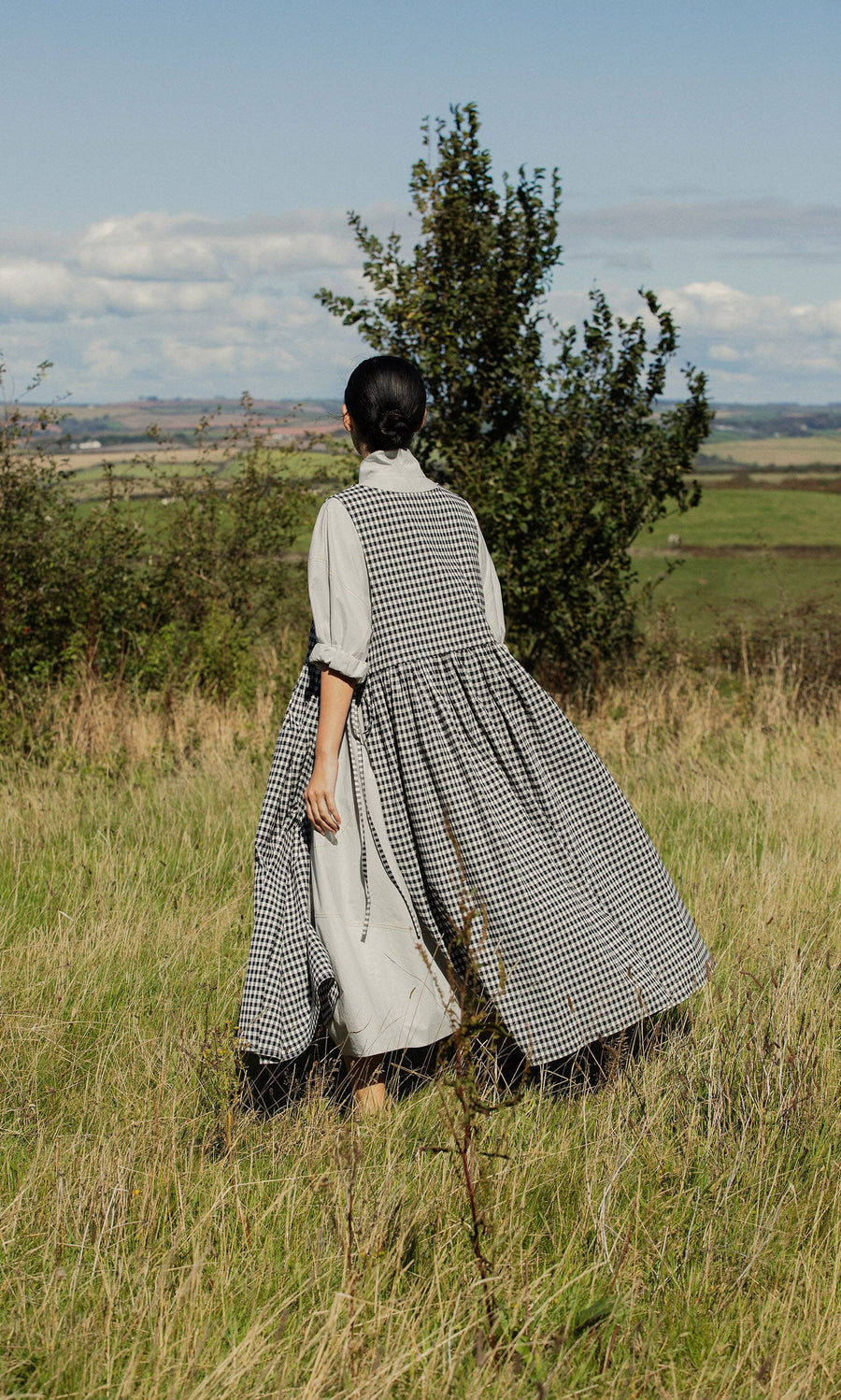Plümo- In a grassy field, someone in the Apron Dress - Gingham, with spacious pockets, gazes toward distant hills.