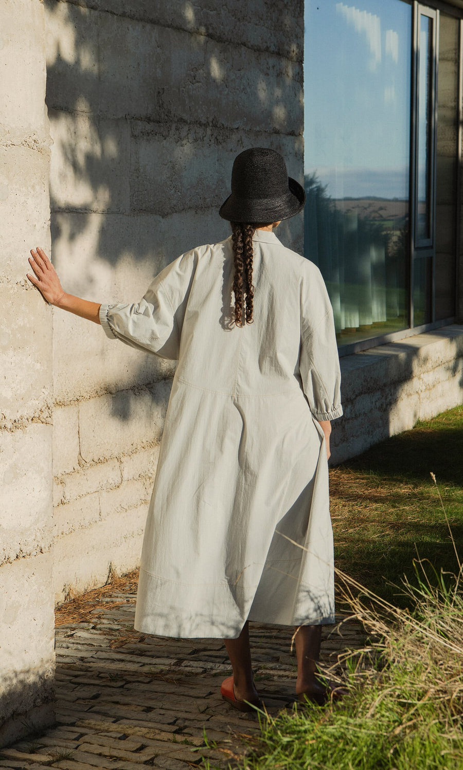 Plümo- Someone wearing the Kaisa Dress with balloon sleeves and a dark hat stands by a sunlit wall, hand touching it.