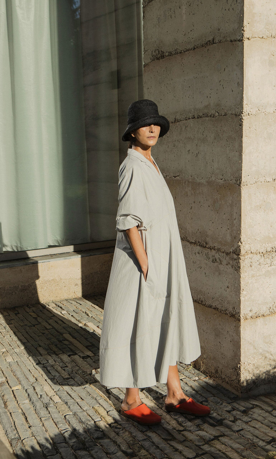 Plümo- Person in a loose Kaisa Dress with balloon sleeves, black hat, and red shoes stands by a concrete building in sunlight.