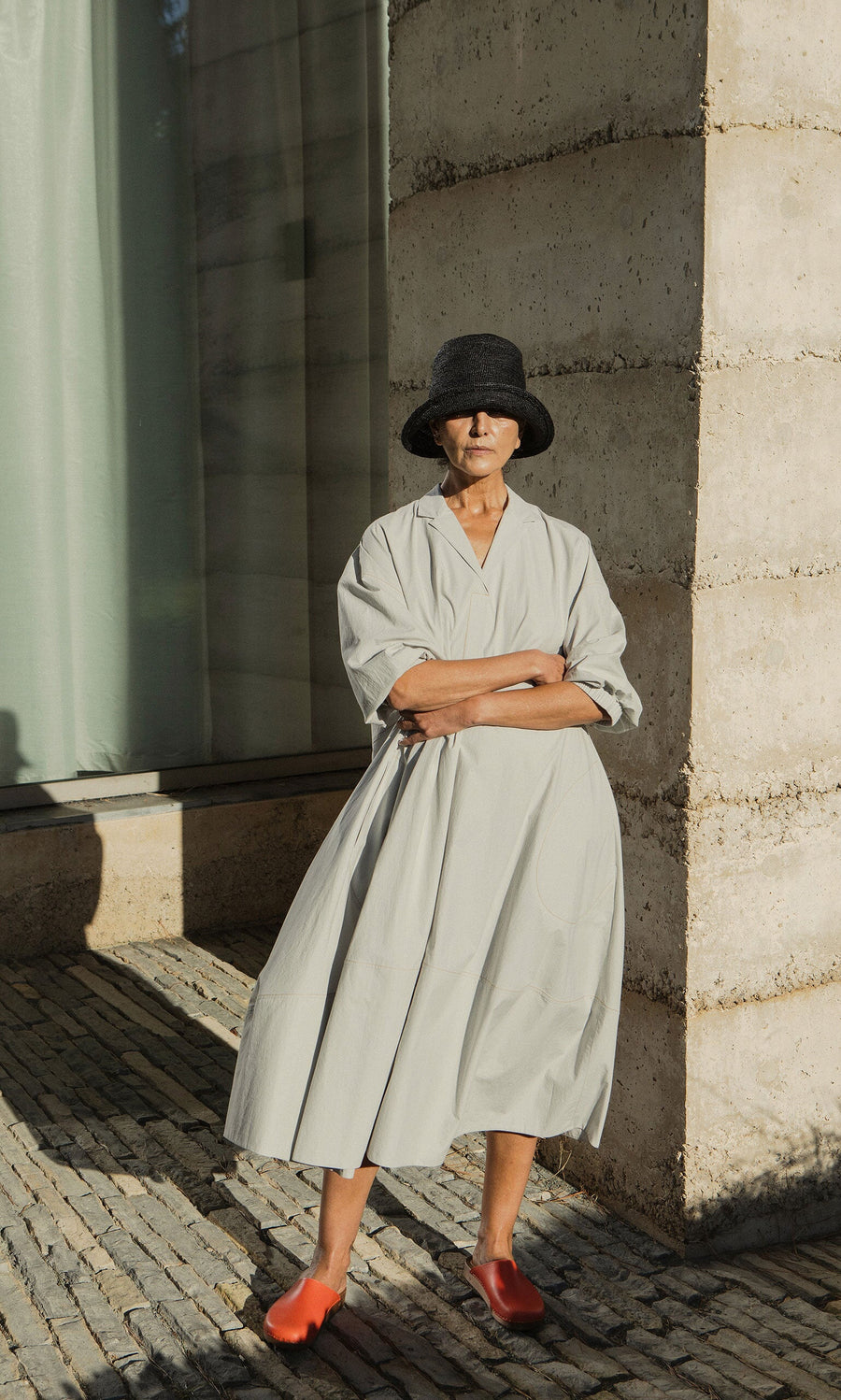 Plümo- Woman in the Kaisa Dress, a wide light gray cotton poplin style with balloon sleeves, stands by a stone wall in sunlight.