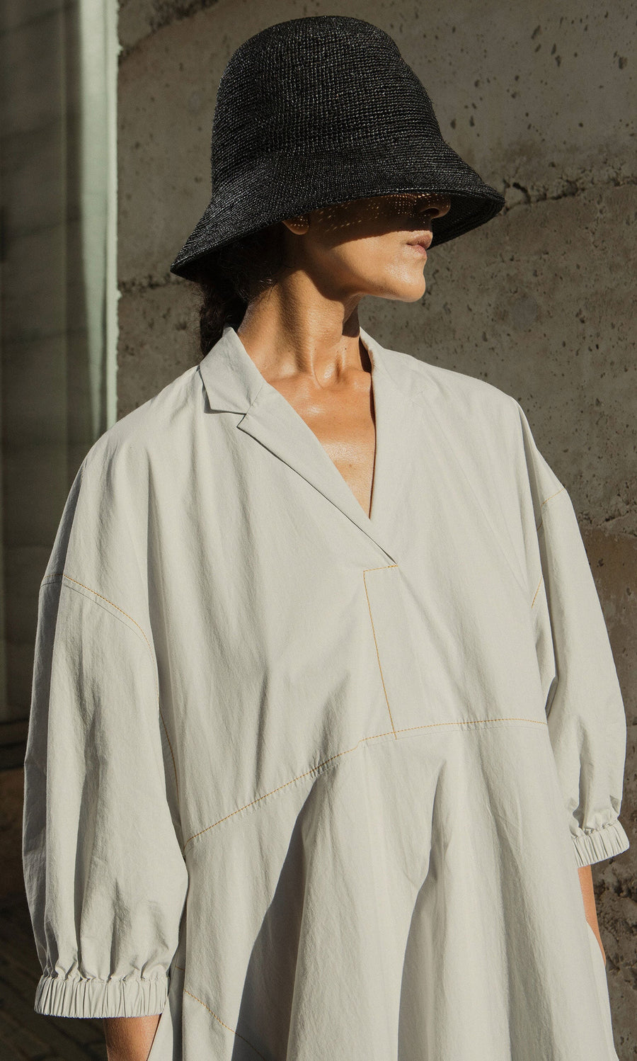Plümo- Person wears the Kaisa Dress with balloon sleeves and a black woven hat, standing against a sunlit concrete wall.