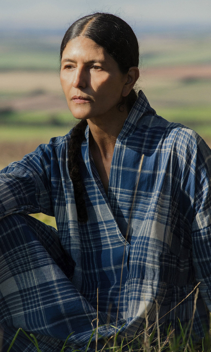 Plümo- Person with long dark hair in a braid wears the Tarkhan Dress - Check, sitting outdoors with fields in the background.