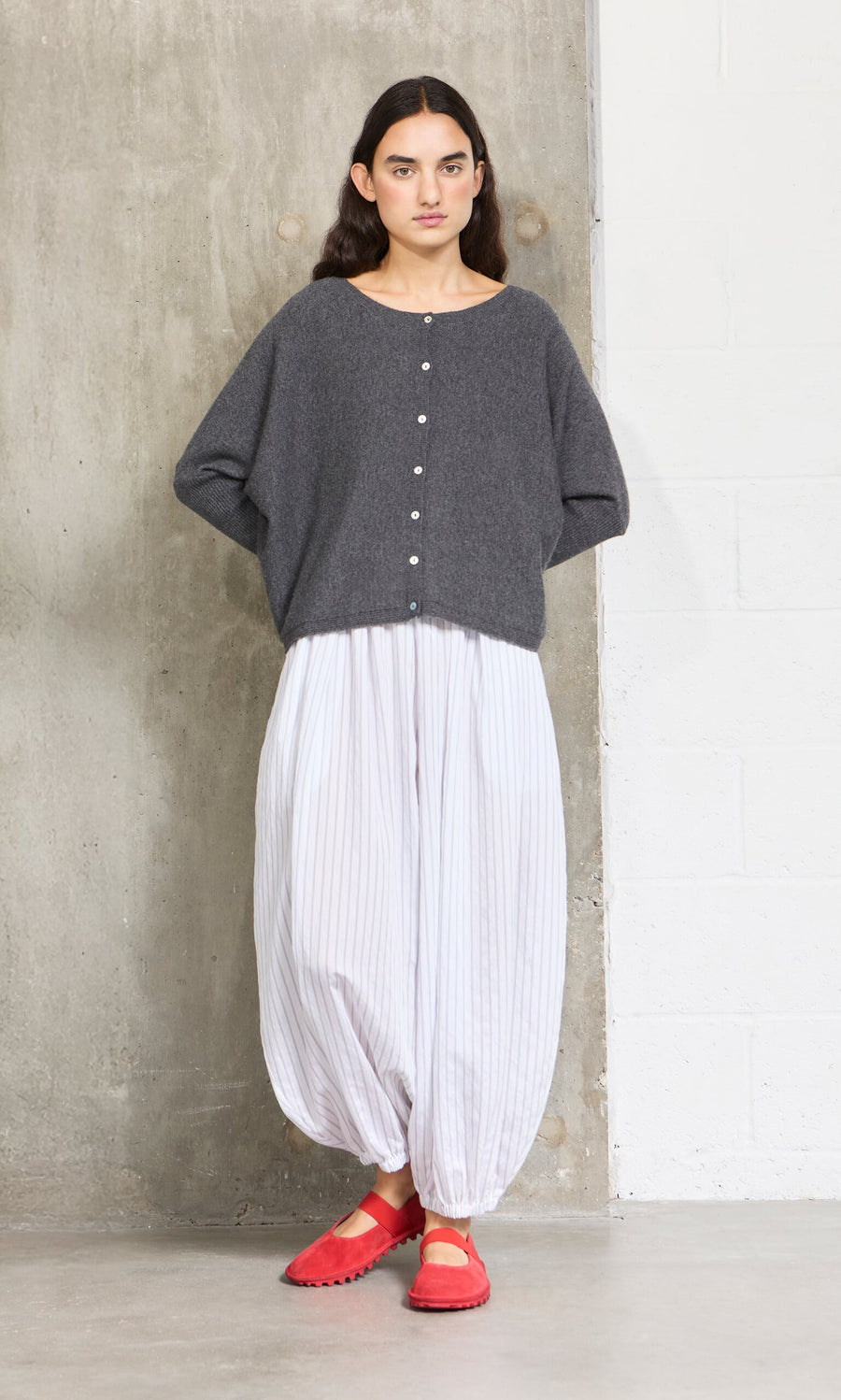 Plümo- A woman in a gray buttoned sweater and white striped pants wears Sola Red Flats by a concrete wall.