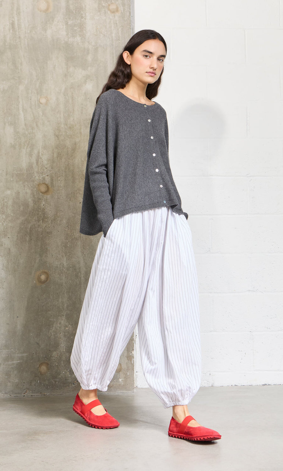 Plümo- Woman in a gray sweater, white striped pants, and Sola Red Flats stands by a concrete and white wall.