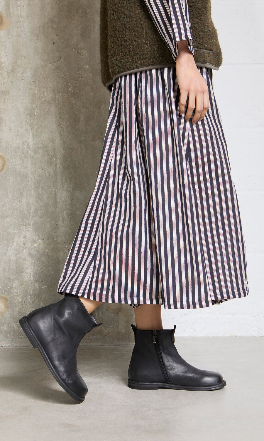 Plümo- Person in a striped dress and Ancona Boots stands on a concrete floor by a textured wall, channeling Danish design.