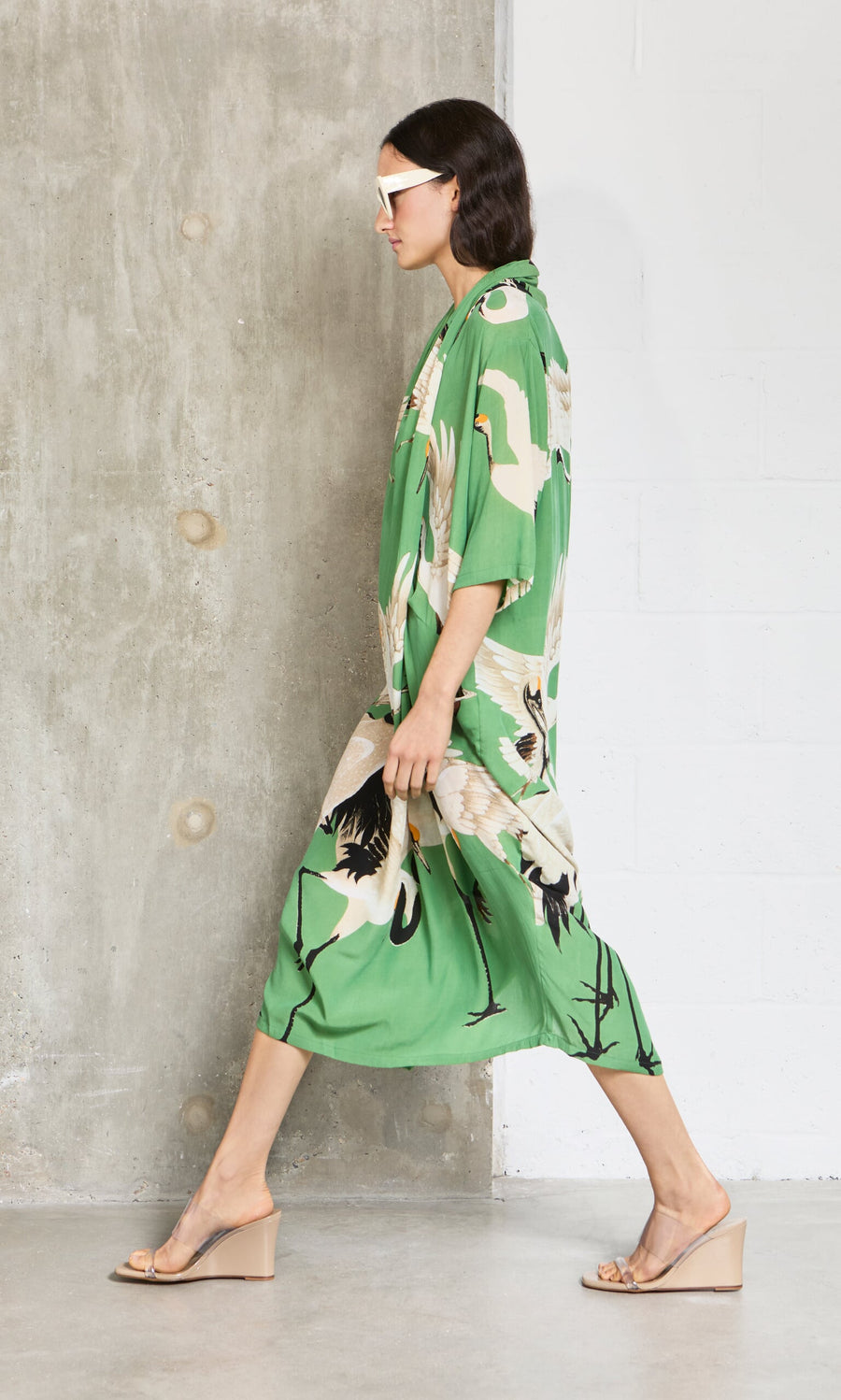 Plümo- Woman in the Green Stork Dress and wedge sandals walks by a concrete wall, sporting stylish white sunglasses.