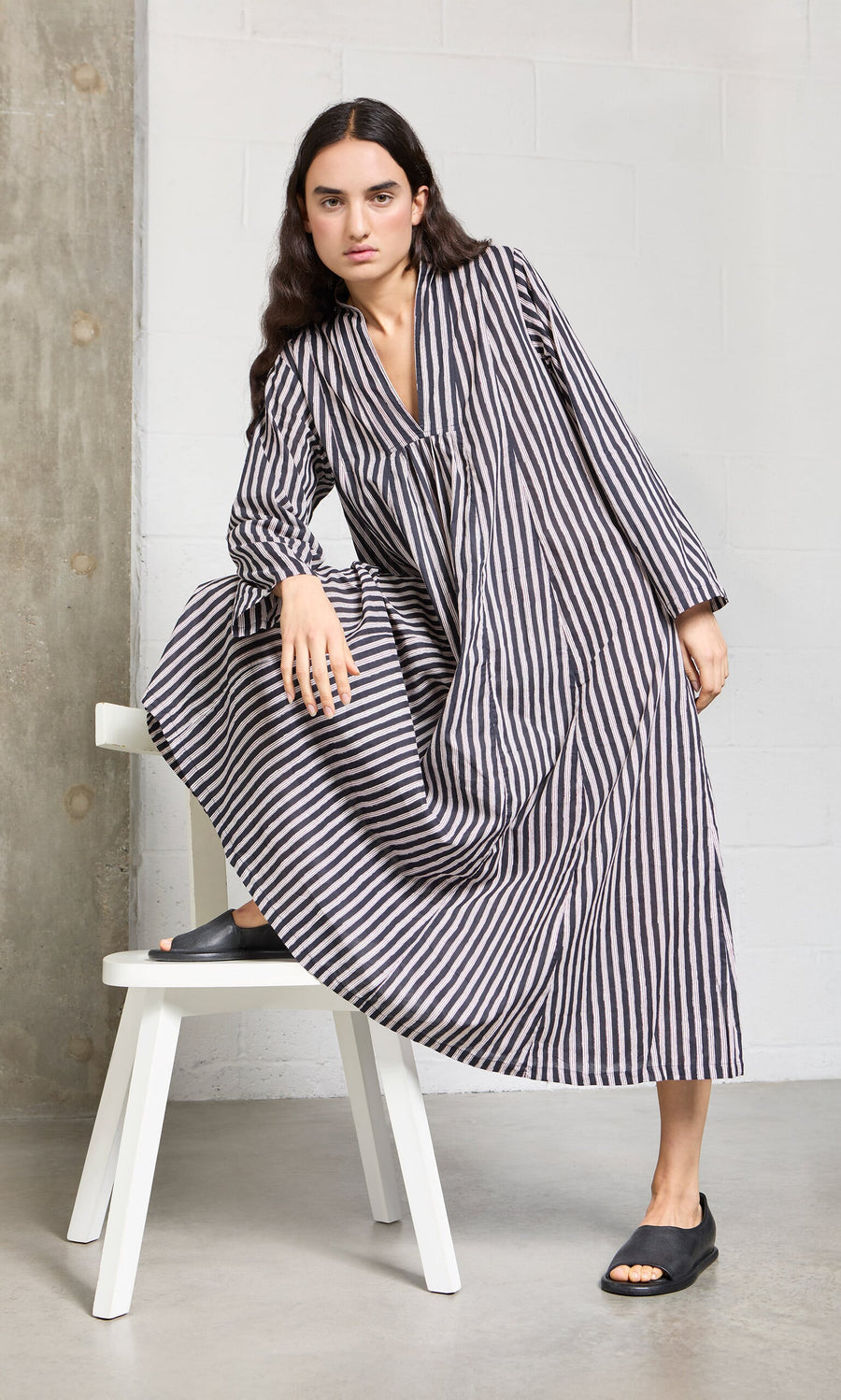 Plümo- Woman in the Hoyland Dress - Charcoal Stripe poses with one knee on a white bench in a minimalistic space.