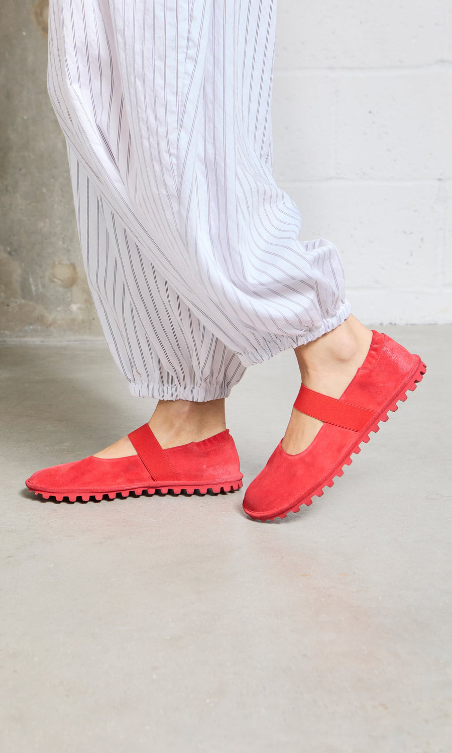 Plümo- Person in loose, white striped pants and Sola Red Flats stands on a gray floor.