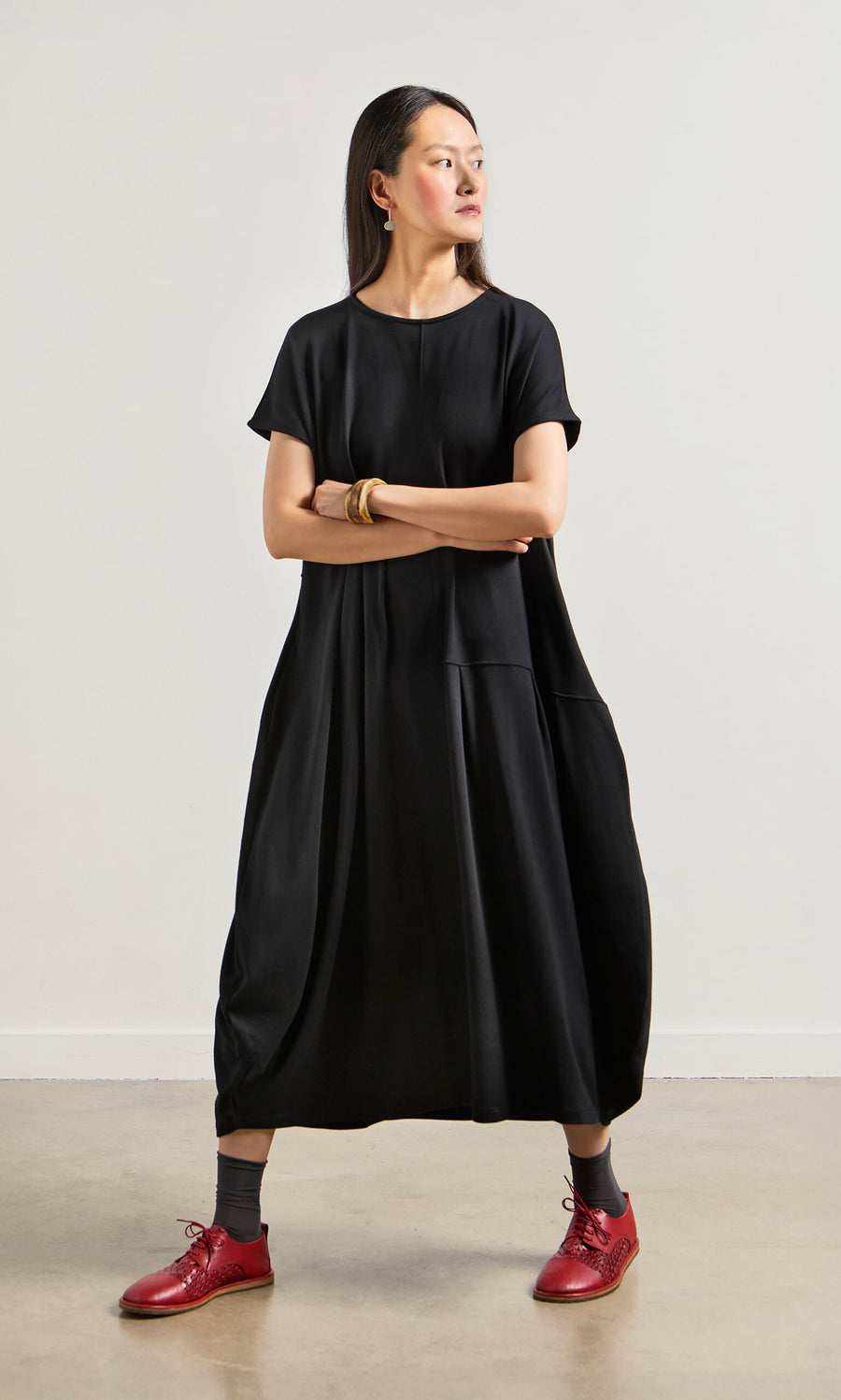 Plümo- Woman in the Blackout Dress stands with arms crossed, wearing red shoes and looking to the side.