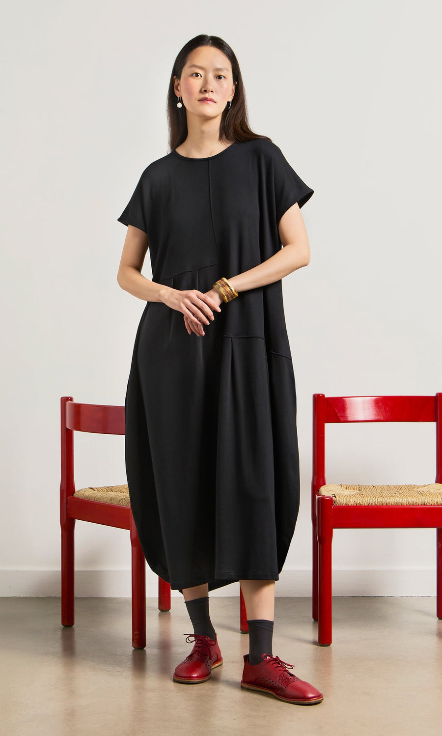 Plümo- Woman in a relaxed fit Blackout Dress and red shoes stands between two red chairs against a white wall.