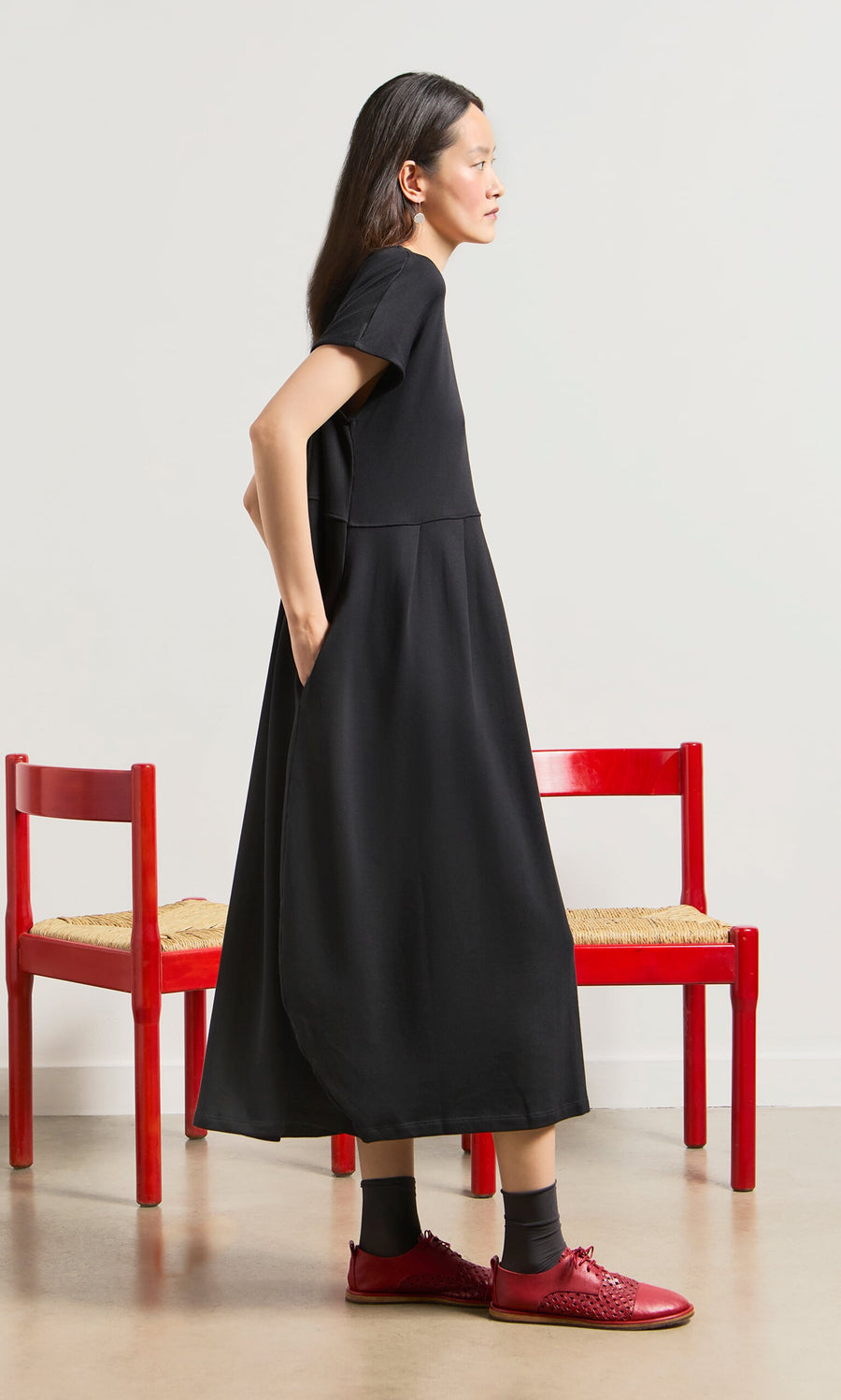 Plümo- Woman in the Blackout Dress and red shoes stands sideways by two red chairs on a neutral background.