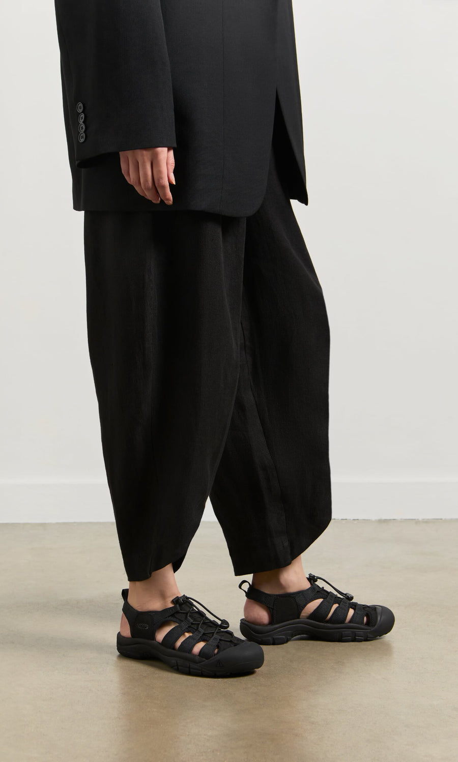 Plümo- A person in loose black pants, a black blazer, and Newport Sandals stands on a concrete floor by a white wall.