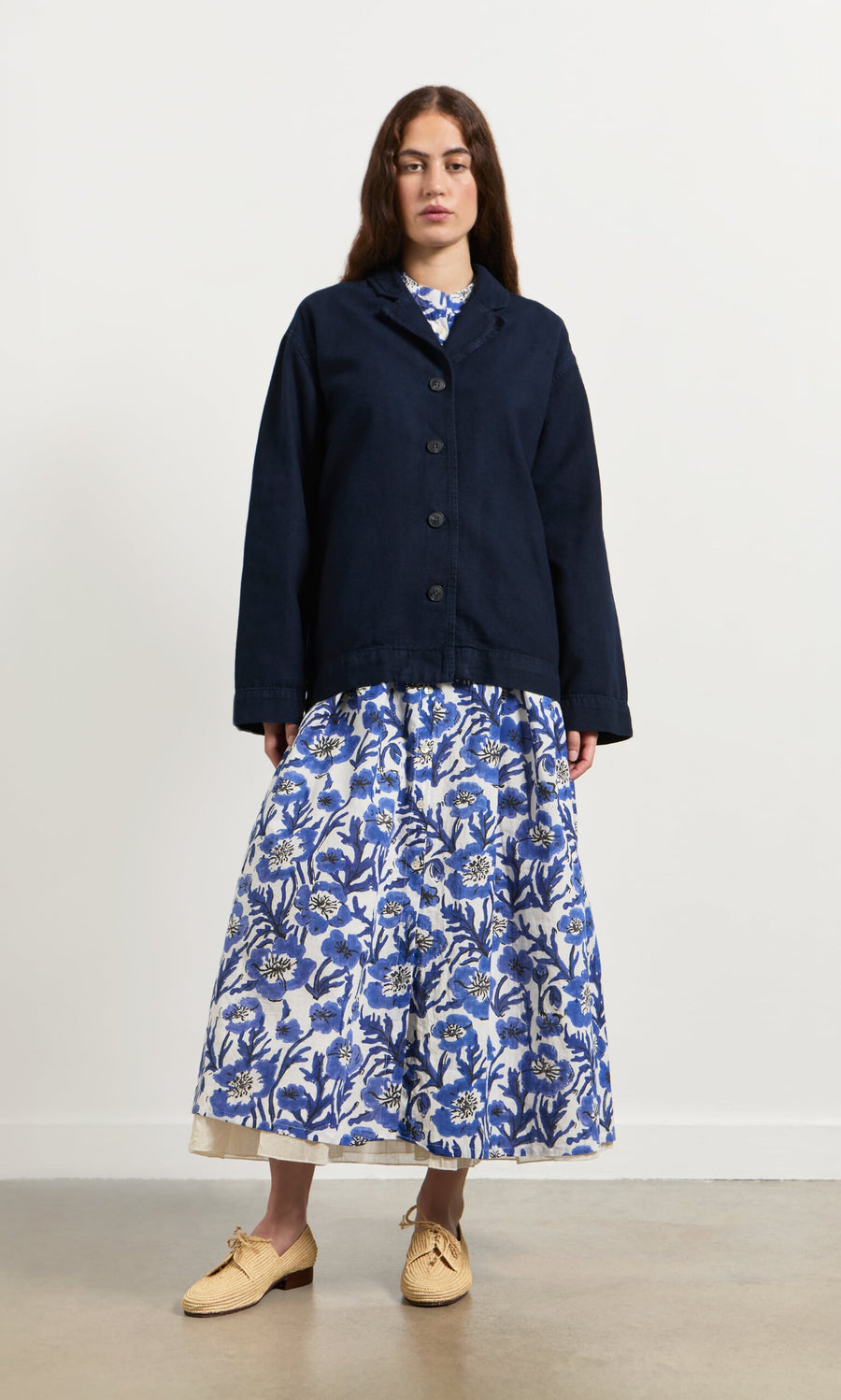 Plümo- Woman in the Blue Pansy Dress with a navy jacket and beige shoes, standing indoors.