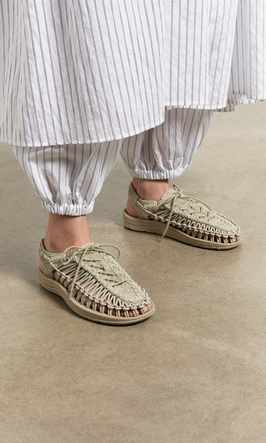 Plümo- Person wearing striped pants, a skirt, and comfy Keen Sandals - Pebble stands on a concrete floor.