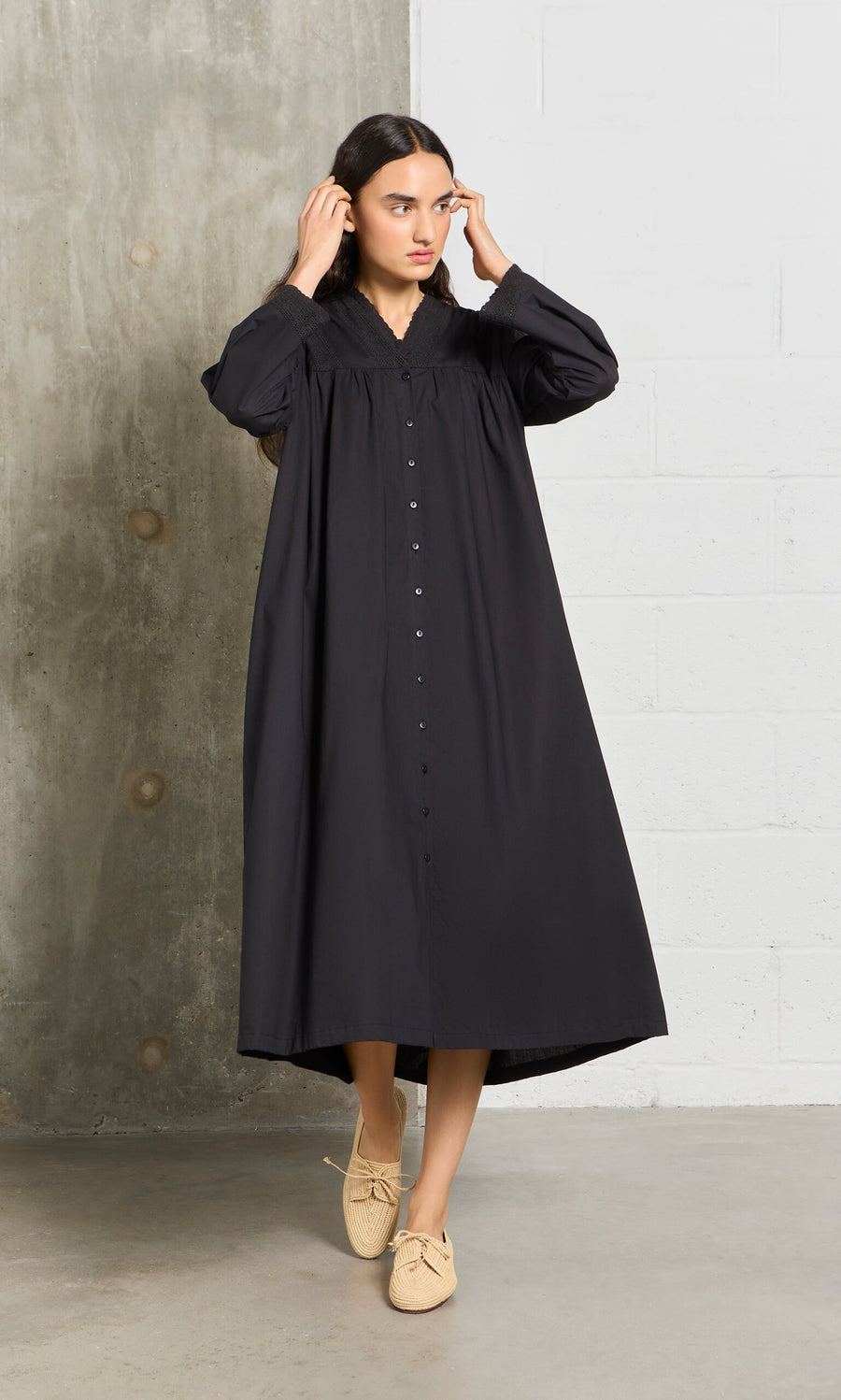 Plümo- Woman wearing the Lily Dress, a long vintage black dress, and tan shoes stands by a concrete and white brick wall indoors.
