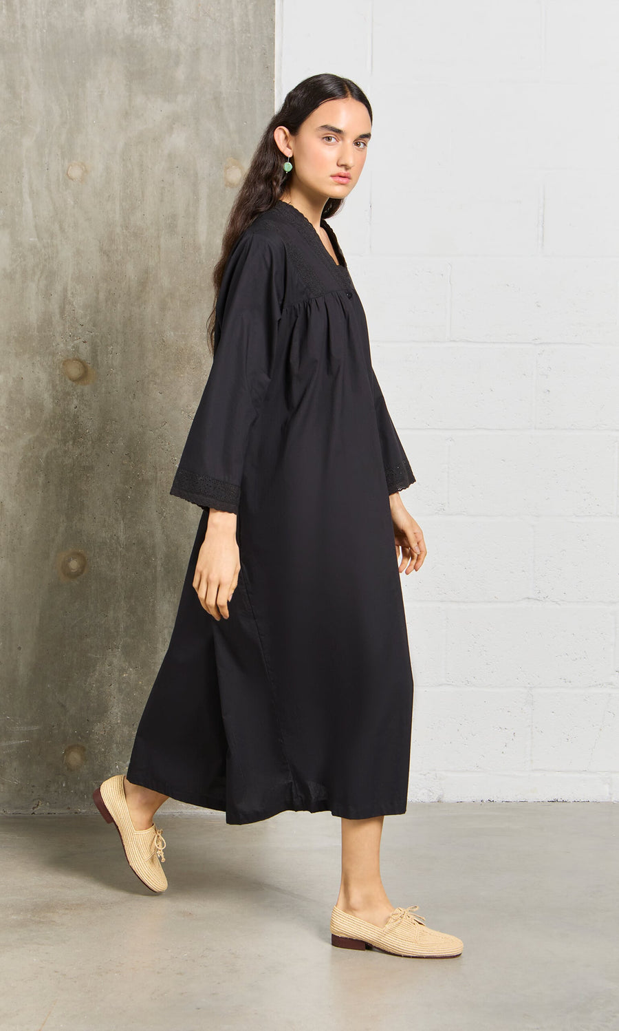 Plümo- Woman wears the Lily Dress and beige shoes, standing indoors by a gray and white wall, looking at the camera.