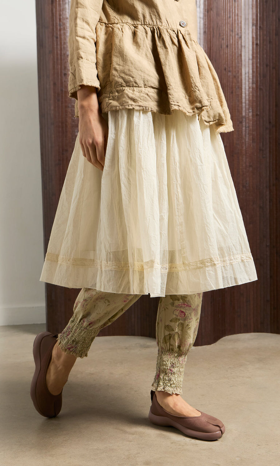 Plümo- Woman stands indoors on a concrete floor wearing Choco Tabi Ballerinas with a beige tunic, white skirt, and floral leggings.