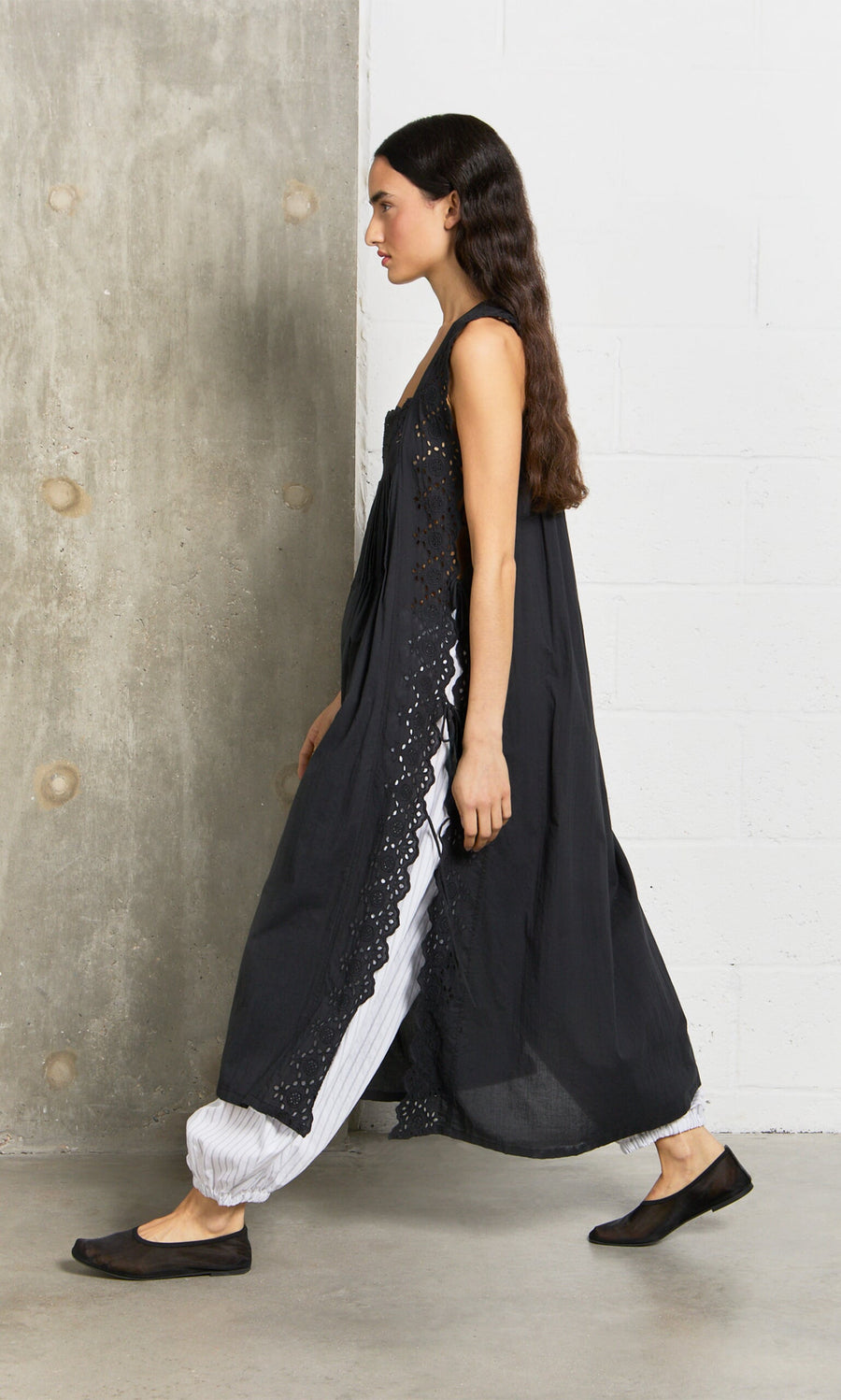 Plümo- Woman in a Vintage Lace Dress over white pants walks by a concrete and white brick wall indoors.