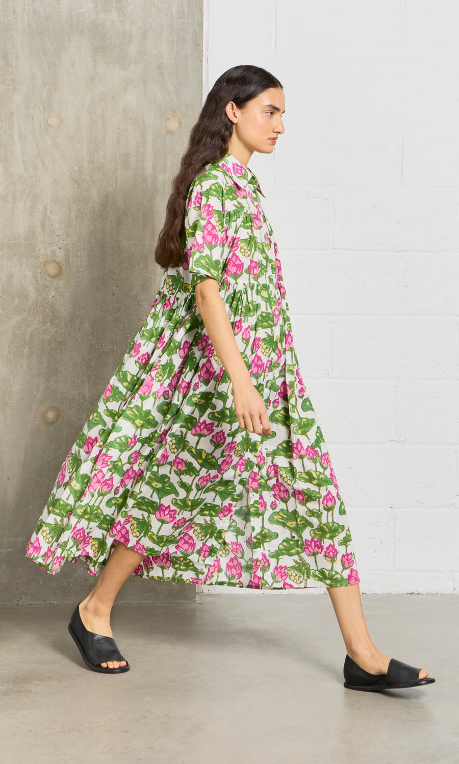 Plümo- A woman in the Waterlily Dress, featuring block-printed cotton voile, walks in black slides by a concrete wall.