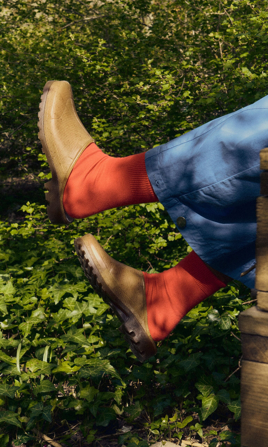 Plümo- Person wearing Opana Mules and bright orange socks raises legs outdoors among leafy greens in blue pants.