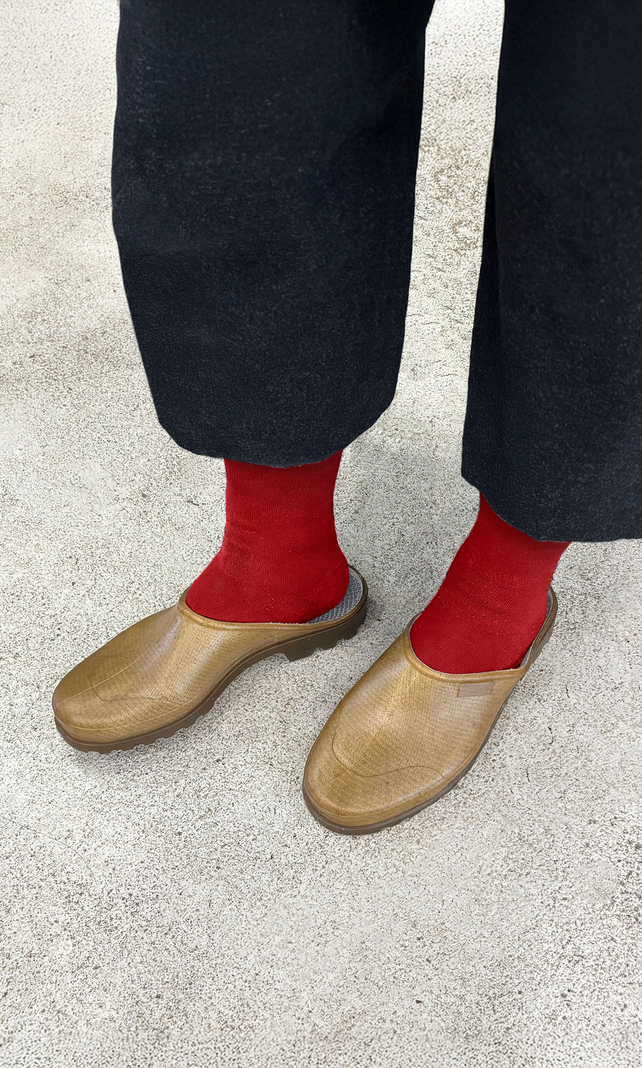 Plümo- Person in black pants wears Opana Mules, brown slip-ons made from recycled plastics, and red socks on a textured surface.